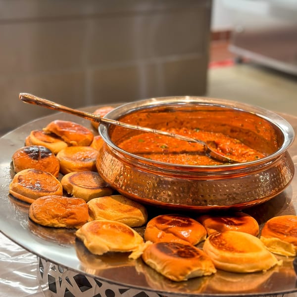 A copper pot of Indian curry with a serving spoon, surrounded by small, golden-brown flatbreads.