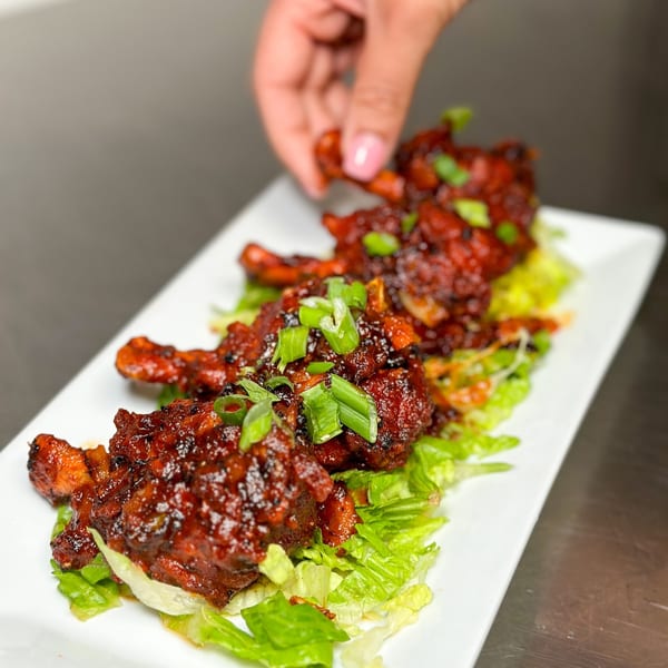 A hand places a piece of Ammrit Indian Cuisine's Chicken Lollipop on a bed of lettuce.